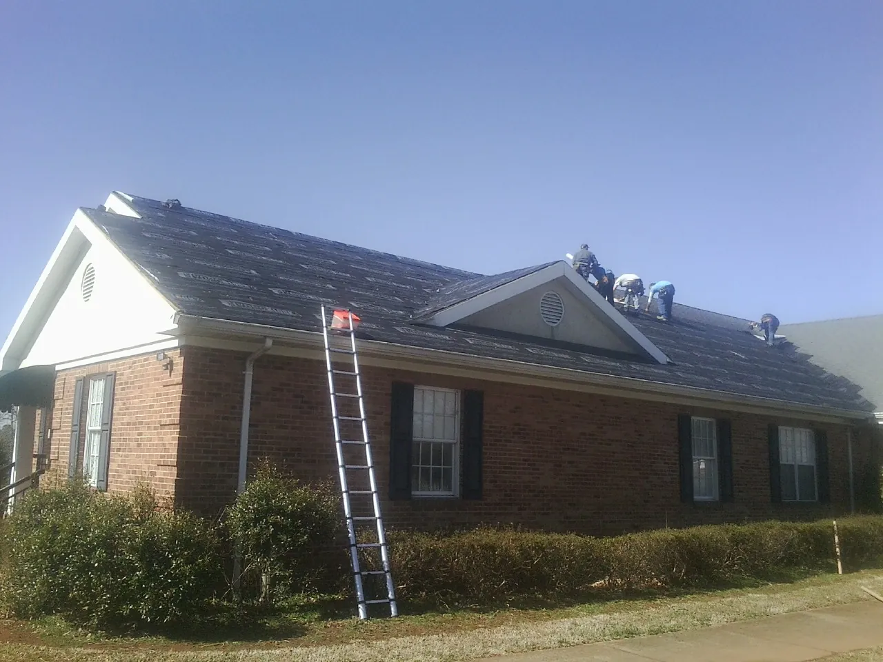 Crew tearing shingles off a brick ranch roof with a ladder set — Emergency Roof Repair work by our crew in Sasser