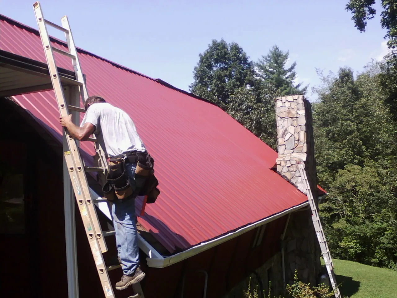 Roofer on a ladder beside a red metal roof and stone chimney — Soffit & Fascia Repair by Roofing Brothers in Sasser